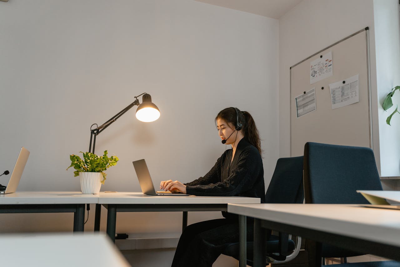 A woman in a modern office setting, wearing a headset and working on a laptop under a desk lamp.